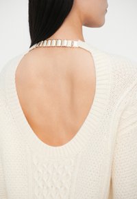 Maglione color crema con schiena scoperta, caratterizzato da un motivo di maglia testurizzato e una tracolla con dettagli dorati nel collo.