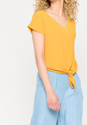 Vrouw met een geel topje met korte mouwen, gestrikt in de taille, en lichtblauwe broek met hoge taille, met krullend blond haar.