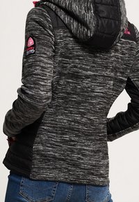 Veste à capuche noire et grise chinée avec des accents matelassés noirs sur les épaules et un détail de logo sur le haut du bras. Présente une coupe ajustée et une texture douce.