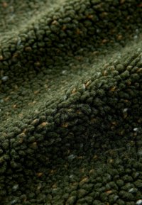 Textured green fabric featuring a soft, fuzzy surface with small, scattered light brown spots. The fabric has a wavy, draped appearance.
