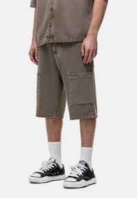 Shorts en denim beige arrivant au genou, dotés de deux grandes poches latérales, associés à des baskets noires et des chaussettes blanches. Finition texturée et coutures visibles.