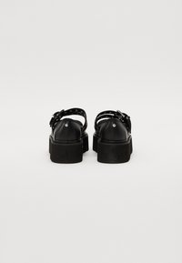 Sandalias de plataforma negras con un upper sintético suave, tiras ajustables en el tobillo y una suela de goma texturizada. La vista trasera muestra un diseño de dos tiras.