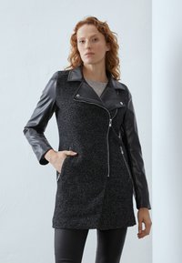 Cappotto nero in tweed con maniche in pelle, zip asimmetrica, due tasche laterali e colletto. Presenta un motivo testurizzato e un design aderente.