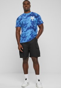 Blå tie-dye kortermet T-skjorte med en hvit logo, kombinert med svarte cargoshorts. Modellen har på seg hvite joggesko og ankelsokker.