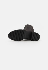 Suela de bota negra de goma texturizada con ranuras horizontales y forma redondeada. Logo de la marca claro presente cerca del talón.