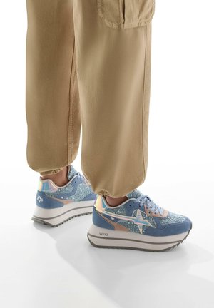 Light blue sneakers with textured surfaces, reflective accents, and a thick white midsole. Worn with khaki tapered pants.