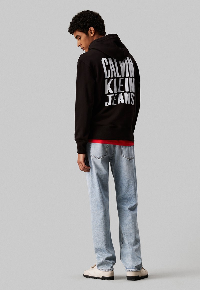 Felpa nera con grande logo bianco "CALVIN KLEIN JEANS" sulla schiena, abbinata a jeans azzurri. Il modello si trova di lato, mettendo in evidenza la vestibilità dell'outfit.
