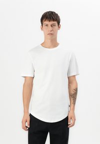Camiseta blanca de algodón con mangas cortas, escote redondo y dobladillo curvado. Combinada con pantalones negros, presenta un tatuaje visible en un brazo.