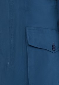 Marinblå jacka med en främre ficka med knappstängning, slät yta och synliga sömnadsdetaljer längs sömmarna.