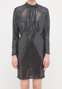 Robe noire à manches longues avec un col haut, un corps froncé et une texture semblable à du tulle ornée de pois métalliques argentés.