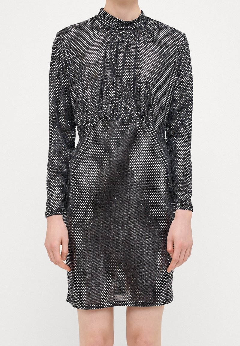 Robe noire à manches longues avec un col haut, un corps froncé et une texture semblable à du tulle ornée de pois métalliques argentés.