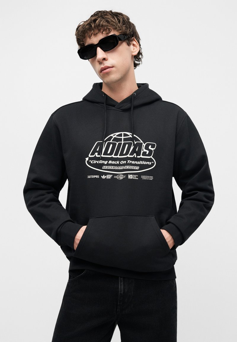 Junger Mann mit lockigem Haar, der einen schwarzen Adidas-Hoodie mit weißem Logo-Text und schwarze Sonnenbrille trägt, Hände in der Kapuzen-Tasche.