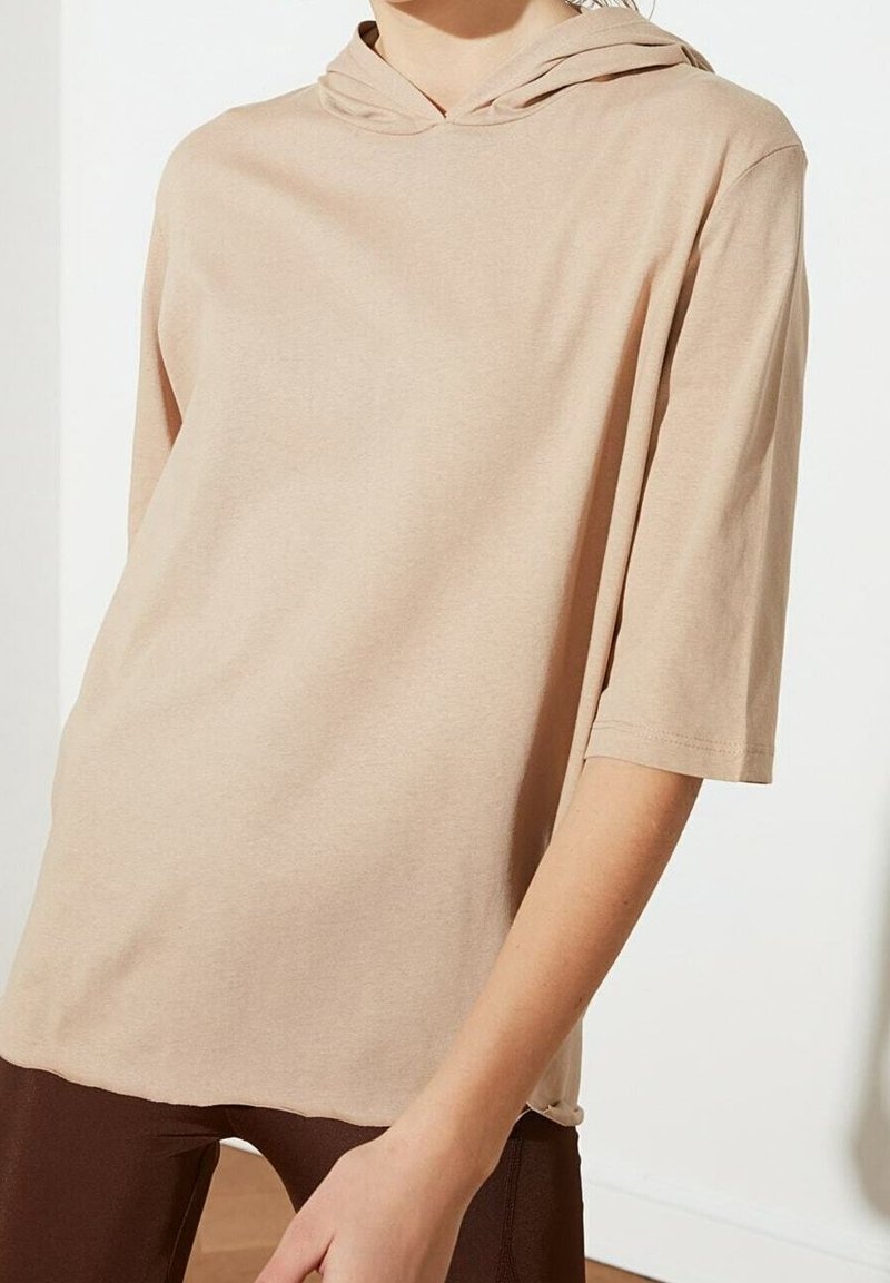 Beige korte mouwen hoodie gedragen over donkerbruine broek, zichtbaar van nek tot halverwege dij, tegen een eenvoudige lichte achtergrond.