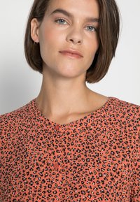 Blusa de estampado de leopardo em coral com fecho de botão. Apresenta um decote redondo e mangas curtas. Textura de tecido suave, estilo casual.