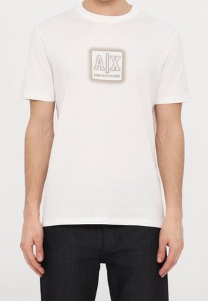 Homme portant un t-shirt blanc Armani Exchange à manches courtes et un pantalon noir, debout devant un fond uni.