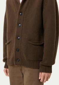 Brun ullkofta med texturerad stickning, med schalkrage, svarta knappar och två framfickor. Ribbstickade ärmslut och nederkant.