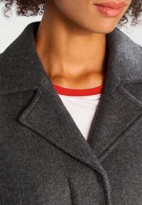 Abrigo de lana gris con cuello vuelto y textura suave. Se lleva sobre una camisa blanca con un detalle rojo en el escote.