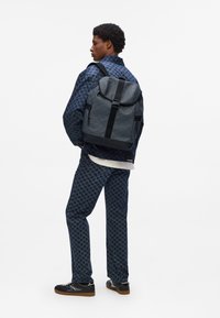 Black patterned backpack with a rounded flap and black straps; worn with a blue houndstooth jacket and matching pants.