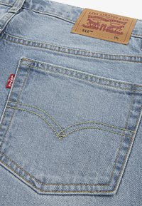 Jean en denim bleu clair avec des coutures jaunes, une poche arrière au design incurvé distinctif et une étiquette en cuir marron à la taille.