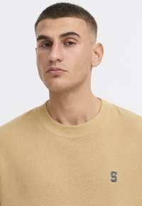 Suéter beige texturizado con un cuello redondo y un pequeño logo bordado de "S" en el pecho. Tejido suave con un patrón de cuadrícula.