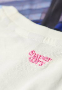 Tejido de algodón crema con una textura suave; presenta un logo bordado en rosa que dice "Super Dry" cerca del escote.