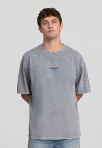 Giovane uomo con capelli corti e ricci, che indossa una maglietta grigia oversized sbiadita con la scritta "Los Angeles" e jeans blu, in piedi contro uno sfondo neutro.