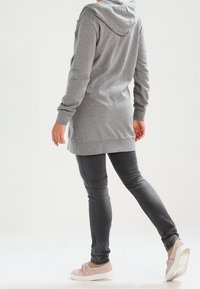 Lungo hoodie grigio con una texture morbida, dotato di cappuccio, tasca frontale a marsupio e polsini a coste, abbinato a jeans slim-fit grigi e scarpe chiare.