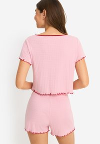 Ροζ σετ δύο τεμαχίων με ριμπ ύφανση, που περιλαμβάνει ένα τοπ με κοντά μανίκια και κόκκινο τελείωμα, και σορτς με βολάν στις άκρες, κατασκευασμένο από μαλακό ελαστικό ύφασμα.