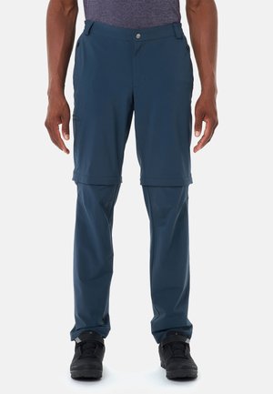 Homme portant un pantalon de randonnée convertible bleu avec short à fermeture éclair et chaussures de randonnée noires, debout devant un fond blanc.