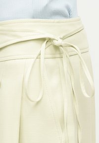 Κοντινή λήψη μιας φούστας σε ανοιχτό μπεζ χρώμα με υφασμάτινη ζώνη δεμένη και πιέτες, φορεμένη με ένα ανοιχτό μπλε ριμπ τοπ.