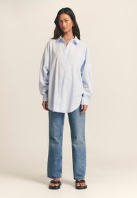 Light blue button-up shirt with long sleeves and a chest pocket, paired with high-waisted blue jeans and black sandals.