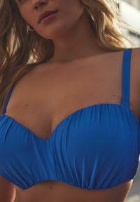 Blauer Bikini-Oberteil aus einem glatten, dehnbaren Material. Mit gerafften Details und dünnen Trägern, die eine strukturierte Form und Unterstützung betonen.