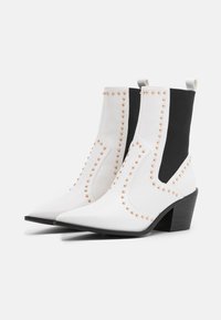 Botas de tobillo de imitación de cuero blancas con puntas afiladas, paneles laterales negros y tachuelas en color oro rosa a lo largo de las costuras. Tacones bajos y gruesos en negro.