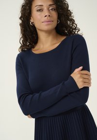 Mörkblå långärmad klänning med rund halsringning, texturerad stickad bodice och plisserad kjol. Anmärkningsvärda egenskaper inkluderar en subtil lyster och figursydda ärmar.