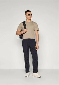 En beige kortermet T-skjorte, marineblå bukser, hvite sneakers, solbriller og en svart ryggsekk bæres av en stående mann mot en nøytral bakgrunn.