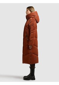 Mujer con abrigo largo acolchado color óxido con capucha, cremalleras y botones a presión, llevando botas negras hasta el tobillo, de pie de perfil sobre un fondo liso.