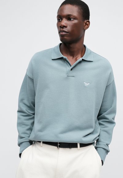 Ljusblå långärmad polo med en vikbar krage, med en enda knapp och en broderad logotyp på bröstet. Bärs med beiga byxor.