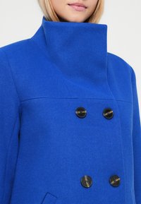 Cappotto di lana blu con colletto alto, design doppiopetto e quattro bottoni neri. Presenta una texture liscia e linee pulite.