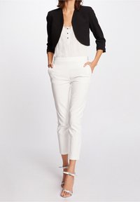 Woman wearing black cropped blazer, white lace-trimmed blouse, white fitted trousers, and silver high-heeled sandals, hands in pockets.