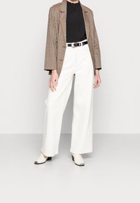Blazer a quadri marrone, top nero arricciato, pantaloni bianchi a gamba larga, cintura nera e stivaletti crema alla caviglia con accenti argentati. Sfondo semplice.