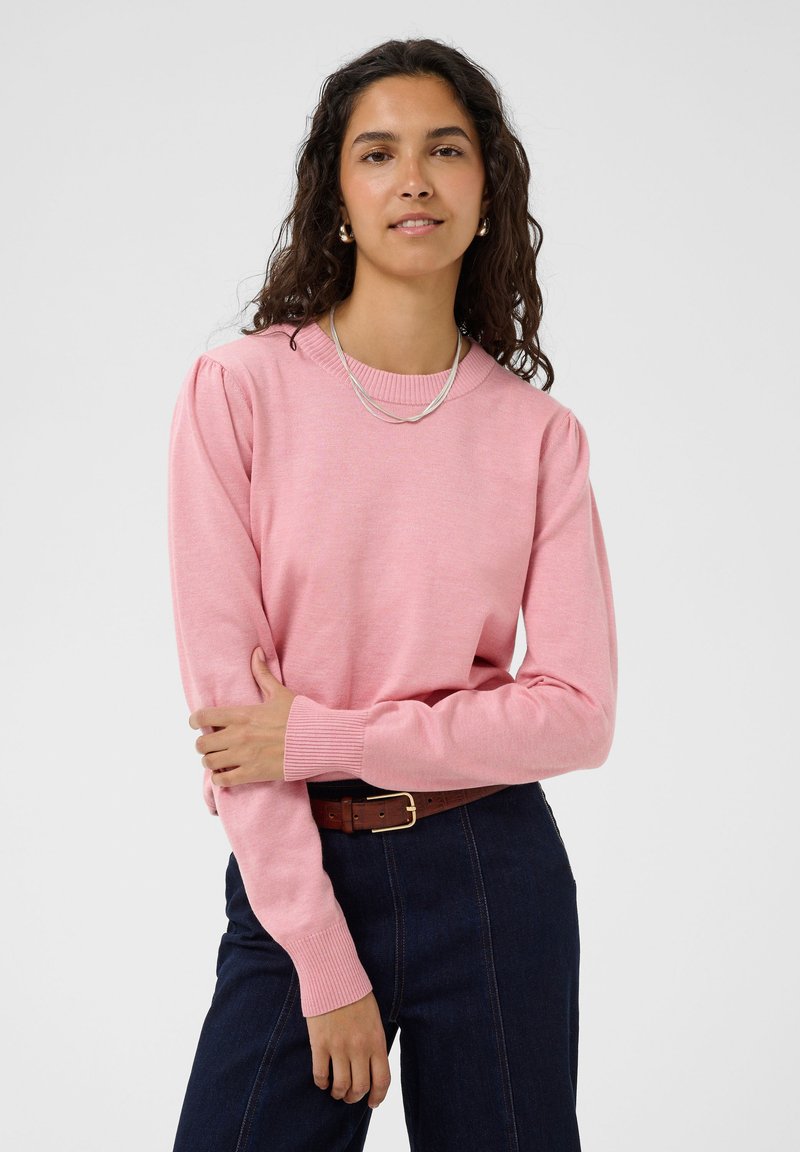 Femme aux longs cheveux bouclés portant un pull rose, un pantalon foncé, une ceinture marron et des bijoux en argent, debout les bras croisés.
