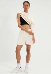 Kremowy sweatshirt z okrągłym dekoltem zarzucony na czarny crop top, w połączeniu z kremowymi szortami. Białe sneakersy z czarnymi akcentami i białe skarpetki. Przezroczyste okulary.