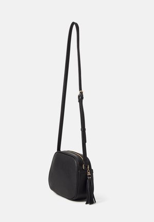 Czarna torebka na ramię z regulowanym paskiem i złotym zamkiem błyskawicznym, ozdobiona czarnym frędzlem na białym tle.