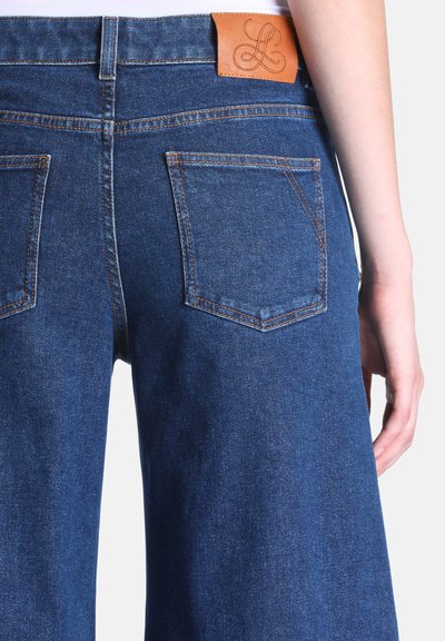 Vue de dos d’une personne portant un jean bleu avec deux poches arrière et un patch en cuir beige sur la ceinture.