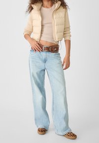 Beżowa kamizelka puchowa na dopasowanej beżowej bluzce, zestawiona z jasnoniebieskimi spodniami o szerokich nogawkach i brązowym paskiem z ćwiekami. Całość uzupełniają klapki w wzór zwierzęcy.