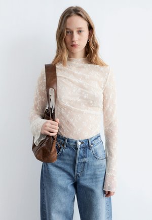 Jeune femme portant un haut transparent à manches longues avec un motif floral, un jean bleu, et un sac à bandoulière en cuir marron, debout devant un fond blanc uni.