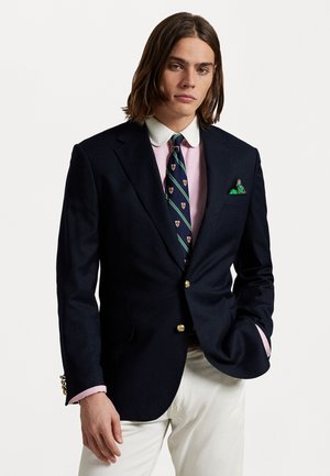 Blazer marine avec boutons dorés, chemise blanche, cravate à motif avec des accents verts et bleus, complétée par un mouchoir de poche vert.
