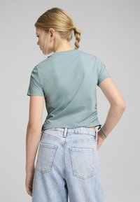 Camisa de manga corta en color teal con un detalle fruncido en el lado, combinada con unos vaqueros de tiro alto de lavado claro que tienen dos bolsillos traseros.