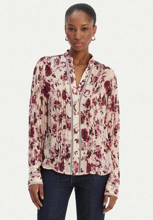 Kobieta ubrana w kremową bluzkę z długim rękawem, z różowym i bordowym kwiatowym wzorem oraz koronkowymi detalami, w połączeniu z ciemnoniebieskimi dżinsami.