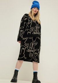 Schwarzes Strickoutfit mit einem Pullover und einem Rock, die abstrakte Gesichtsmuster in Beige zeigen. Das Model trägt eine blaue Mütze und klobige schwarze Stiefel.